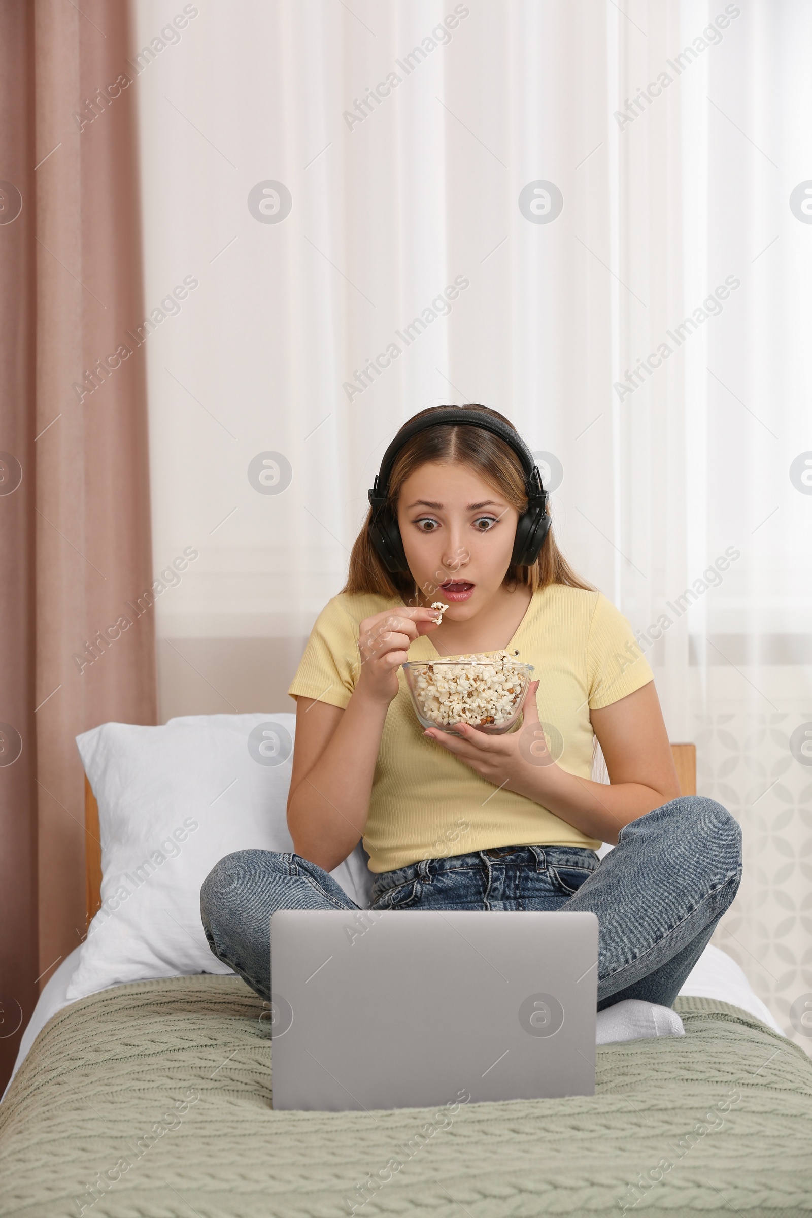 Photo of Teenage girl with headphones eating popcorn while using laptop on bed at home