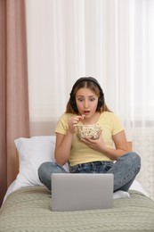 Teenage girl with headphones eating popcorn while using laptop on bed at home