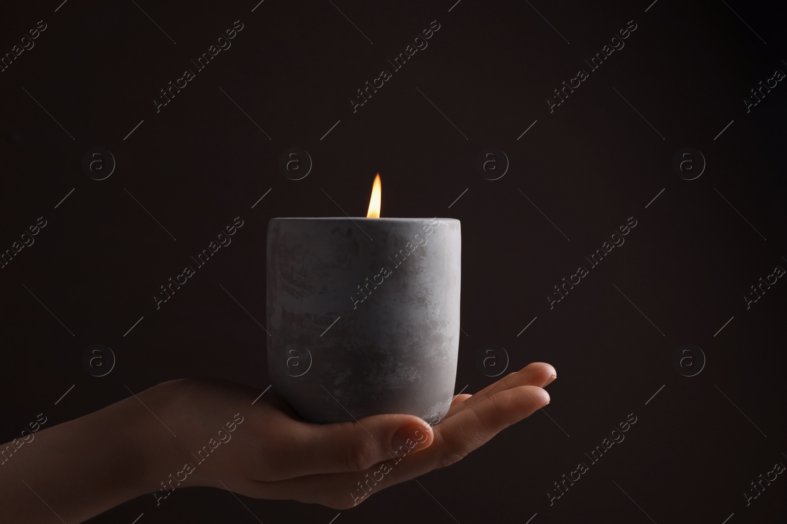 Photo of Woman with lit candle in concrete holder against dark brown background, closeup
