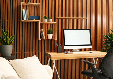 Comfortable workplace with computer near wooden wall in stylish room interior. Home office design