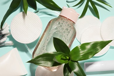 Bottle of micellar cleansing water, cotton pads and green plants on turquoise background, flat lay