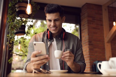 Handsome man with cup of coffee and smartphone at cafe in morning