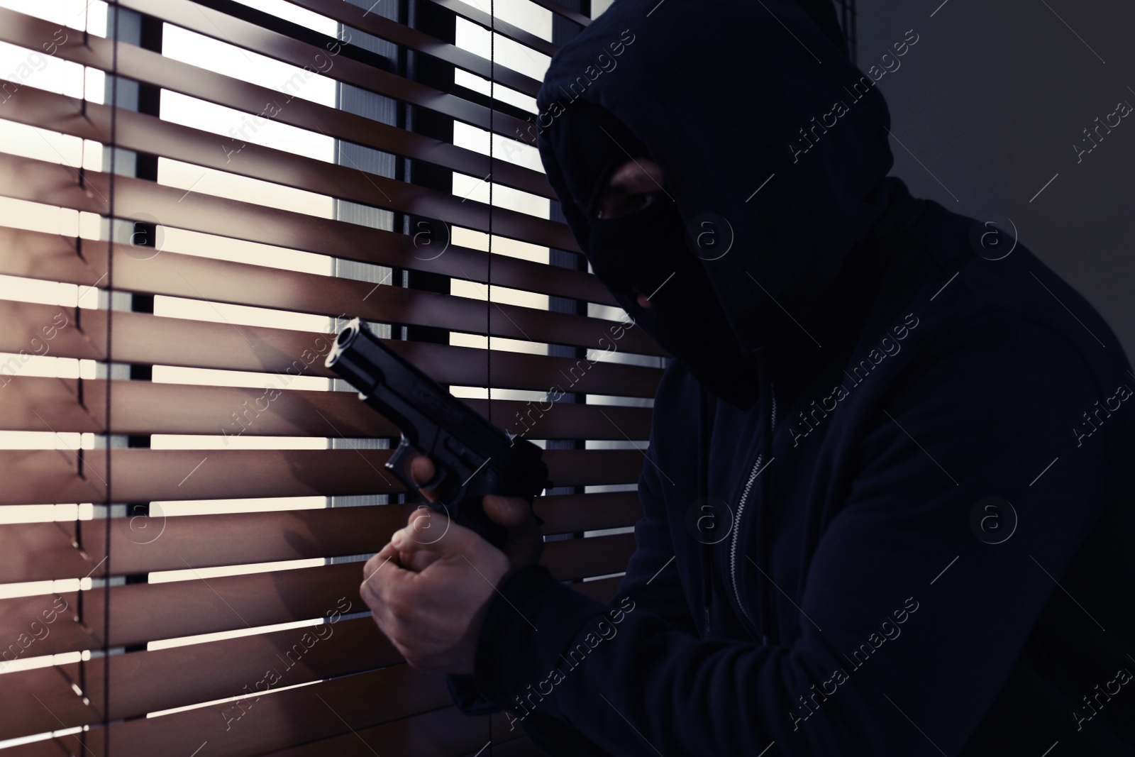Photo of Masked man with gun spying through window blinds indoors. Dangerous criminal