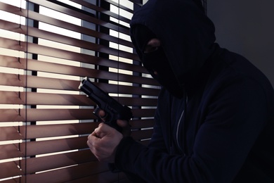 Masked man with gun spying through window blinds indoors. Dangerous criminal