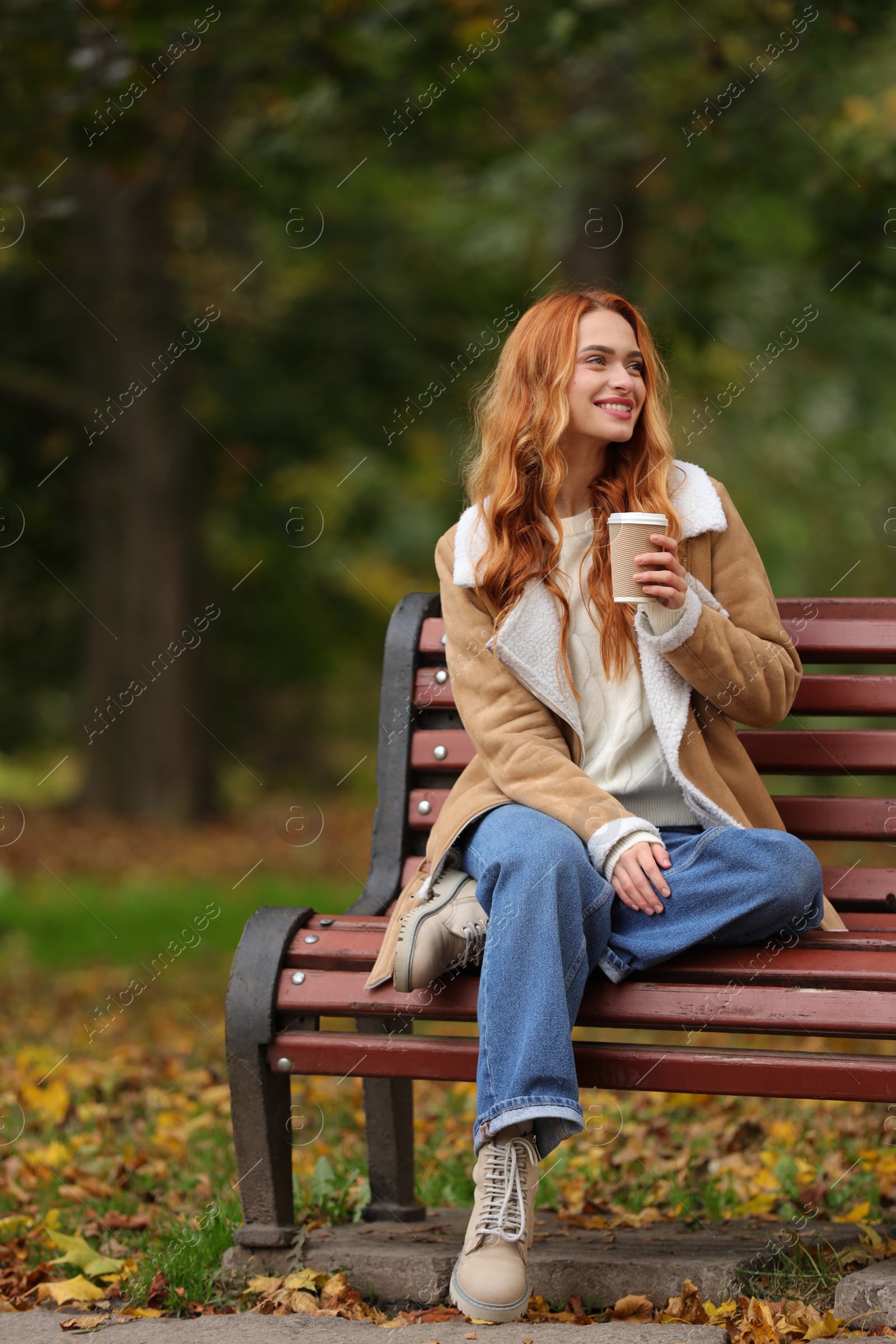 Photo of Smiling woman with paper cup sitting on bench in autumn park