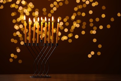 Photo of Hanukkah celebration. Menorah with burning candles on table against brown background with blurred lights, space for text