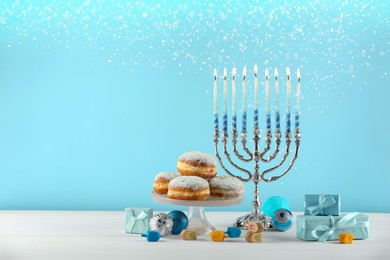 Image of Hanukkah celebration. Menorah with burning candles, dreidels, gift boxes and donuts on white wooden table against light blue background, space for text