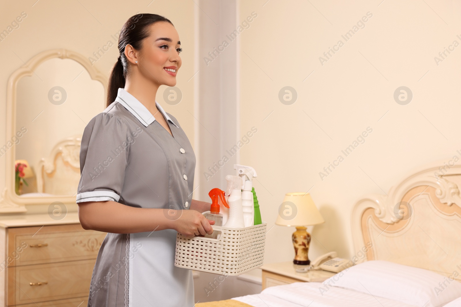 Photo of Young chambermaid holding basket with cleaning products in hotel room. Space for text