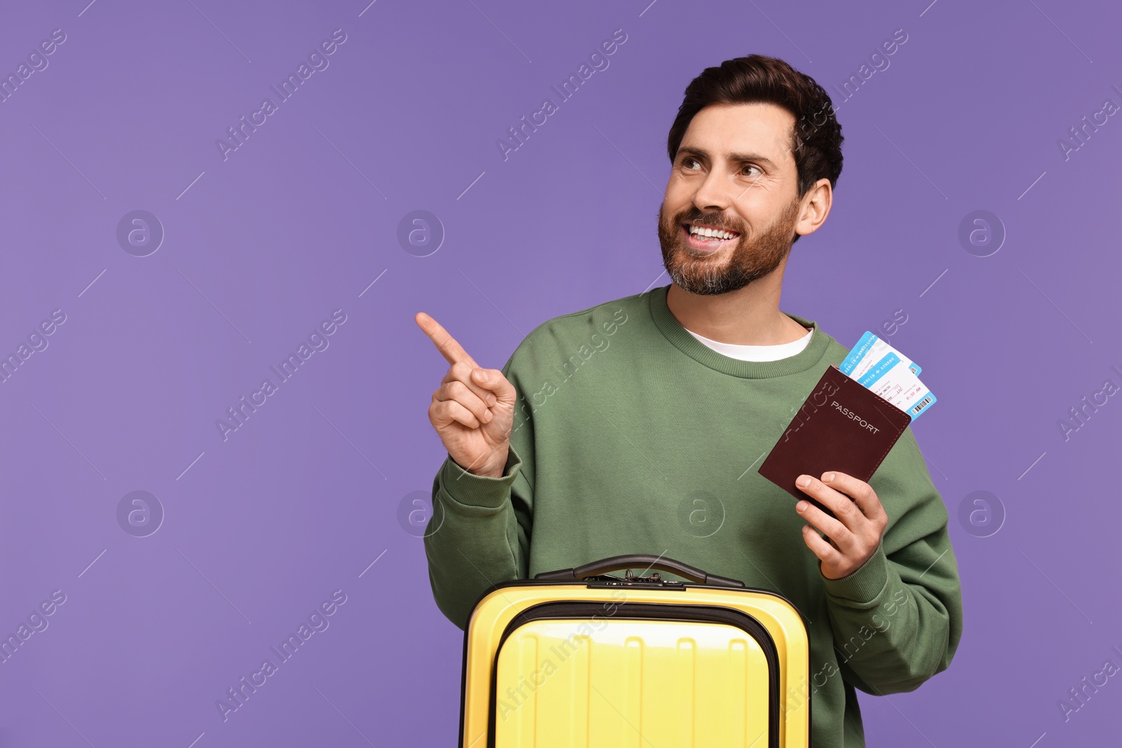 Photo of Smiling man with passport, tickets and suitcase pointing at something on purple background. Space for text