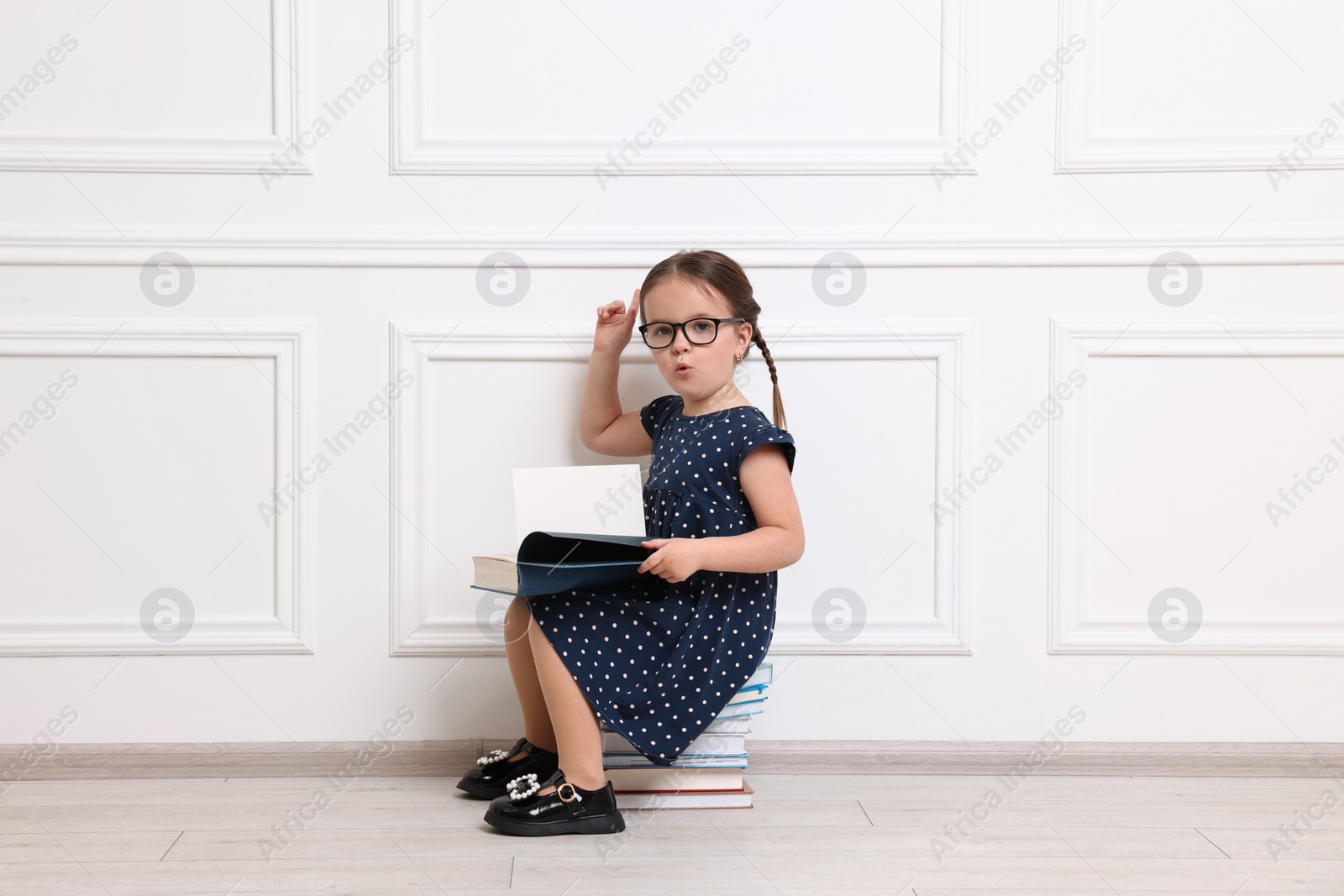 Photo of Cute little girl in glasses sitting on stack of books near white wall