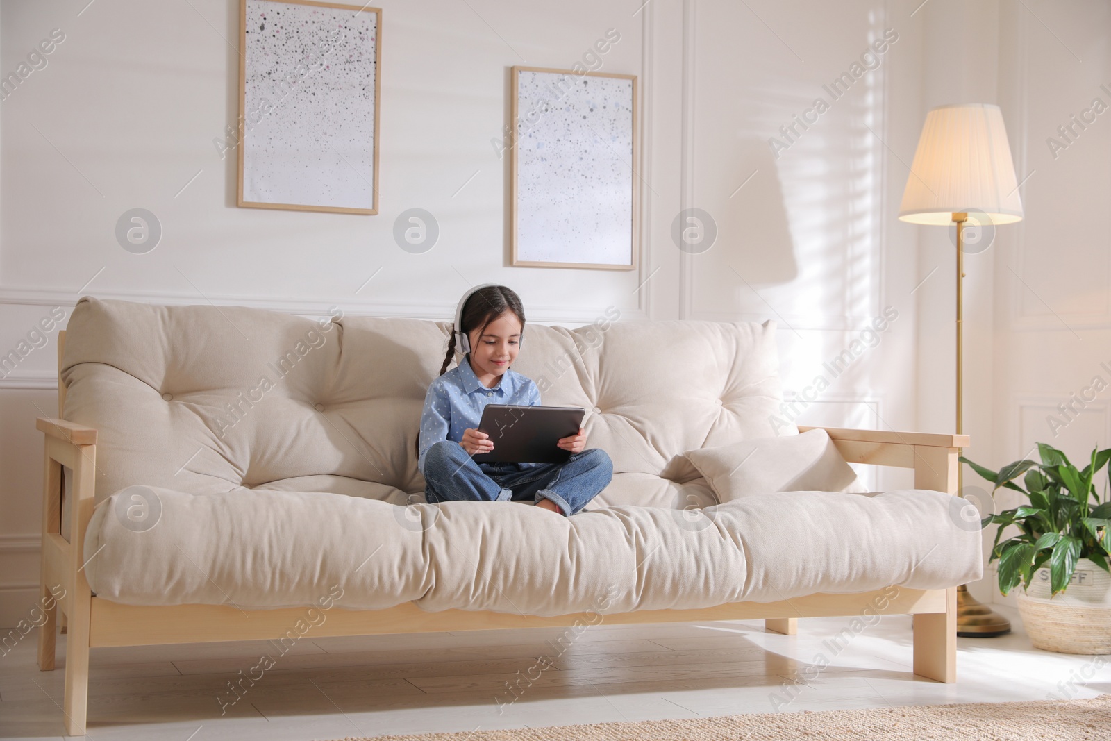 Photo of Little girl with headphones and tablet on sofa at home