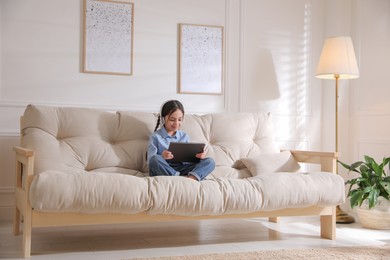 Little girl with headphones and tablet on sofa at home