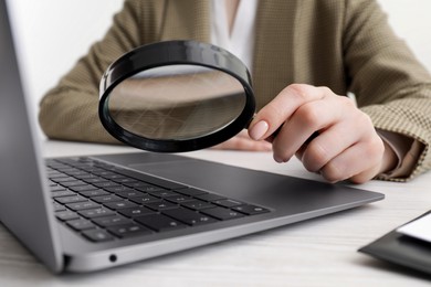 Photo of Woman holding magnifier near laptop at white wooden table, closeup. Online searching concept