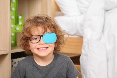 Photo of Happy boy with eye patch on glasses near shelf indoors. Strabismus treatment