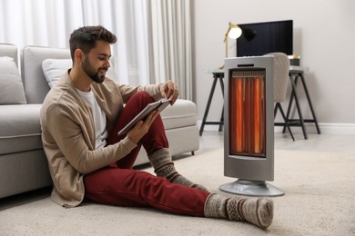 Photo of Young man reading book near electric heater at home
