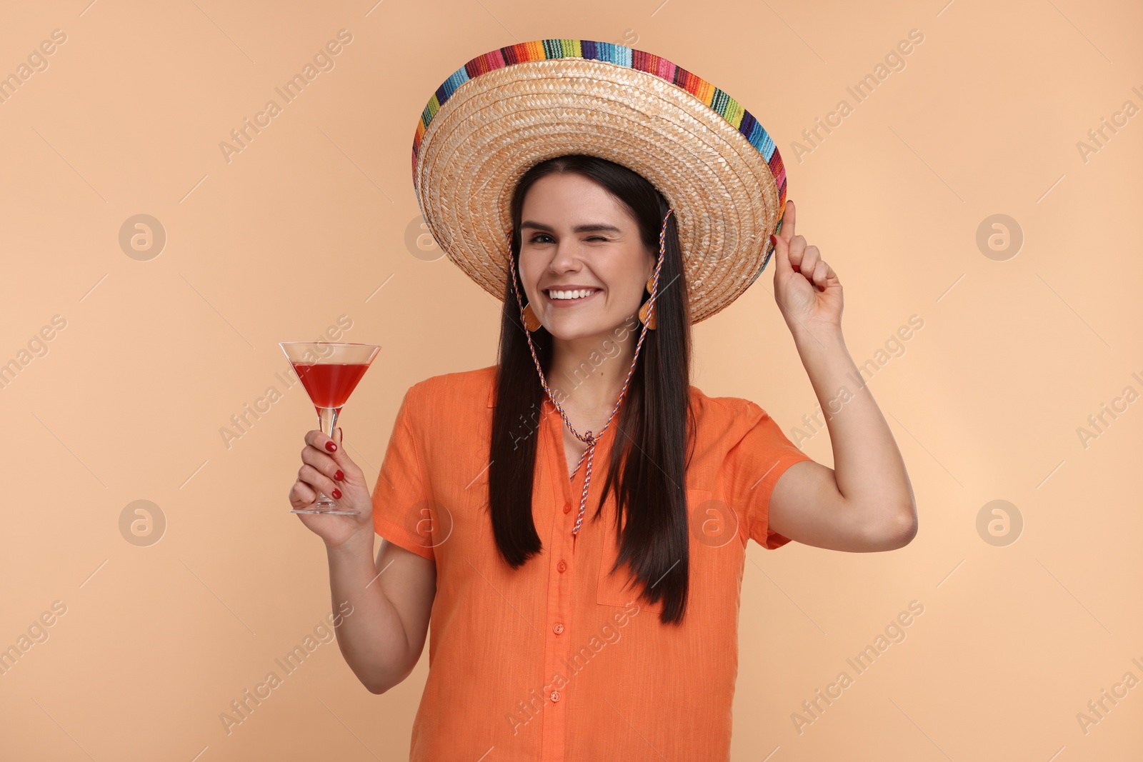 Photo of Young woman in Mexican sombrero hat with cocktail winking on beige background