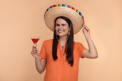 Photo of Young woman in Mexican sombrero hat with cocktail winking on beige background