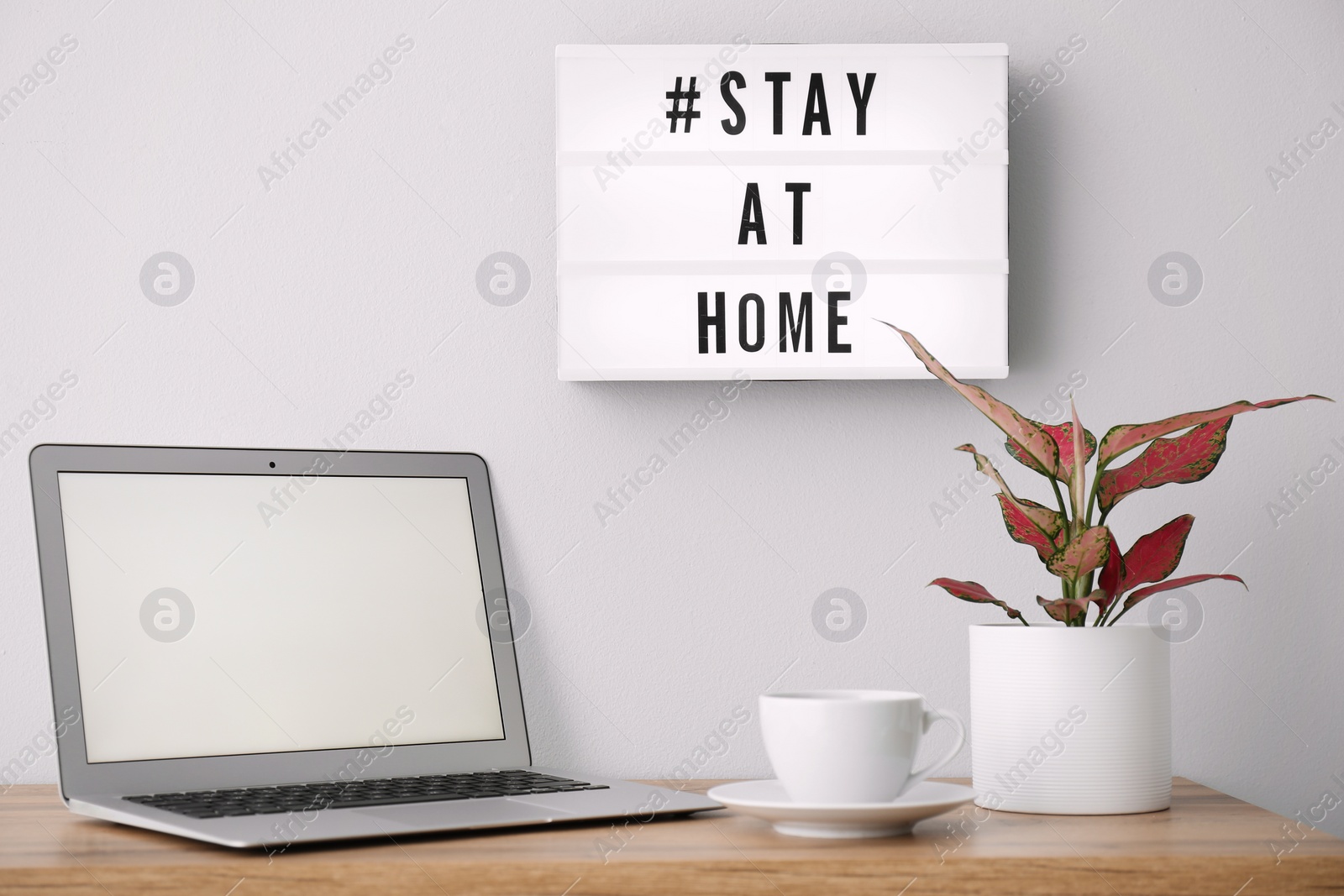 Photo of Laptop, cup and lightbox with hashtag STAY AT HOME indoors. Message to promote self-isolation during COVID‑19 pandemic