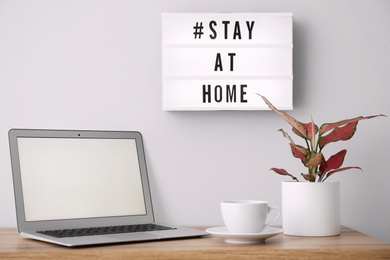 Photo of Laptop, cup and lightbox with hashtag STAY AT HOME indoors. Message to promote self-isolation during COVID‑19 pandemic
