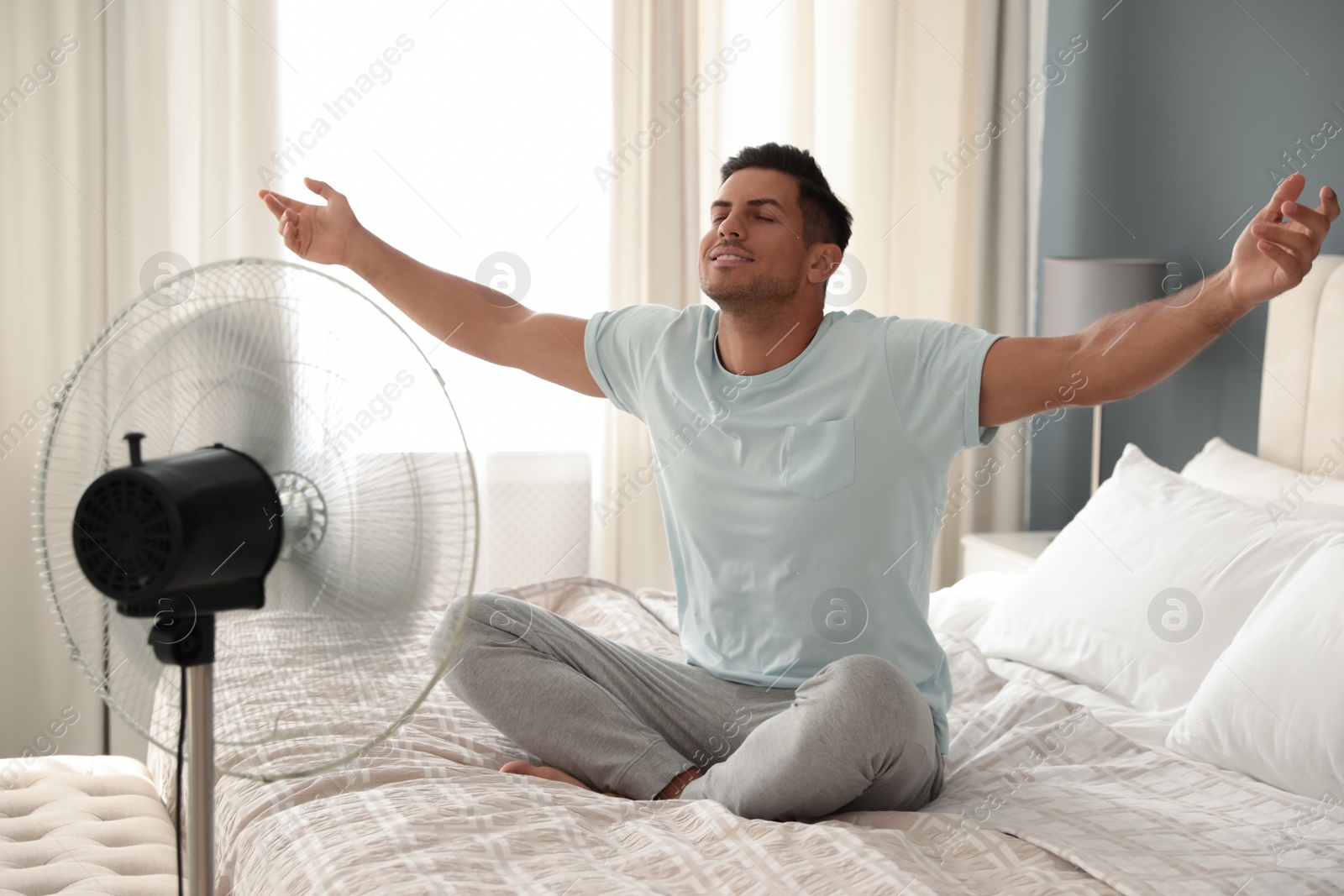 Photo of Man enjoying air flow from fan on bed in room. Summer heat