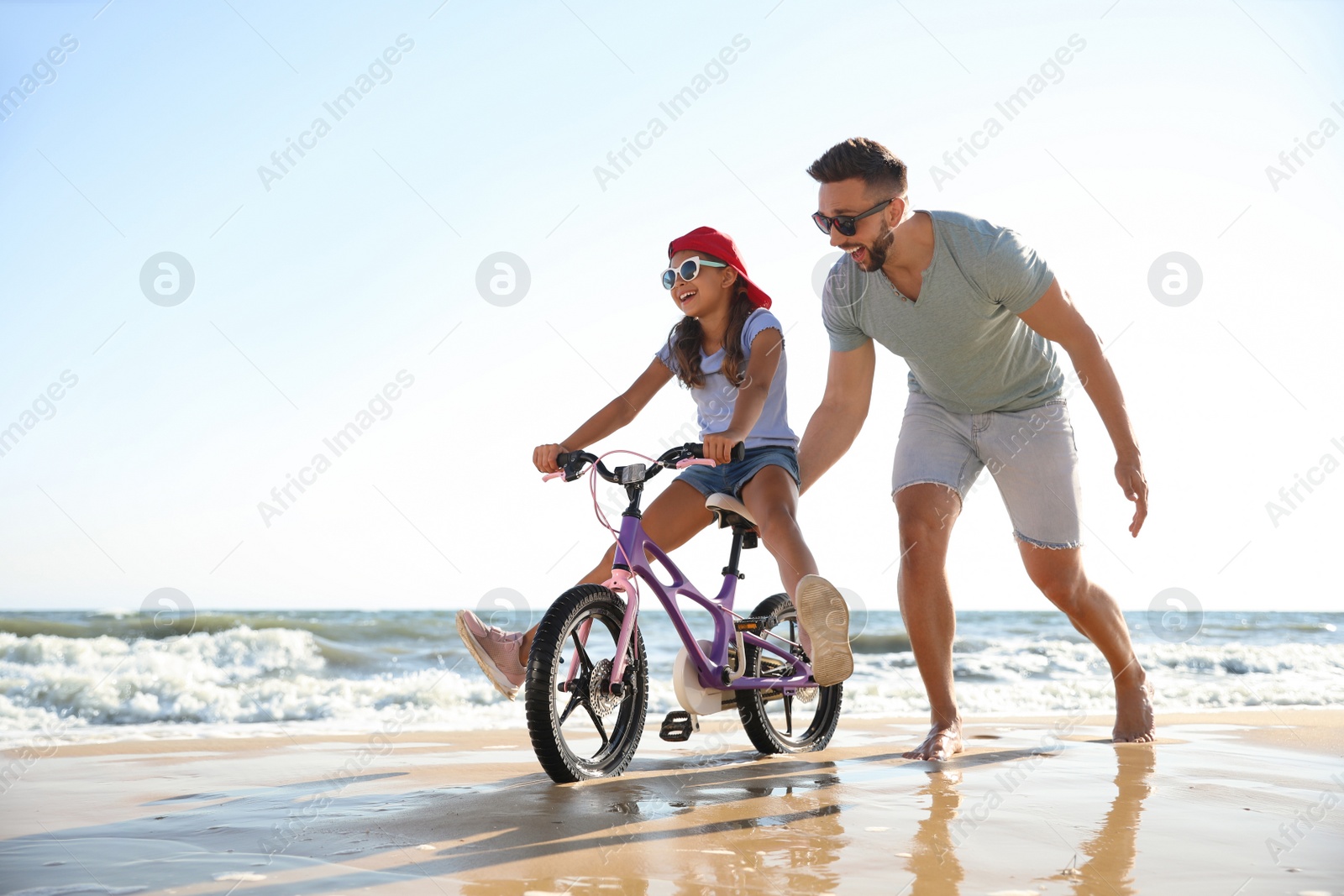 Photo of Happy father teaching daughter to ride bicycle on sandy beach near sea