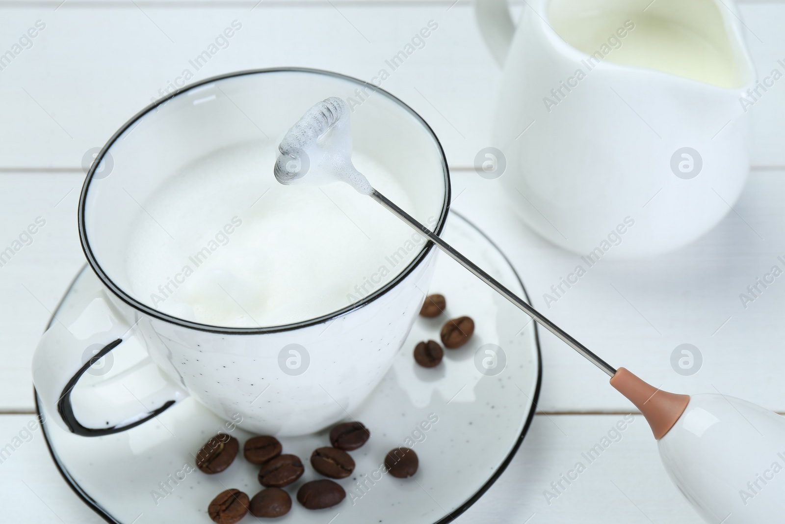 Photo of Mini mixer (milk frother), whipped milk and coffee beans on white wooden table, closeup