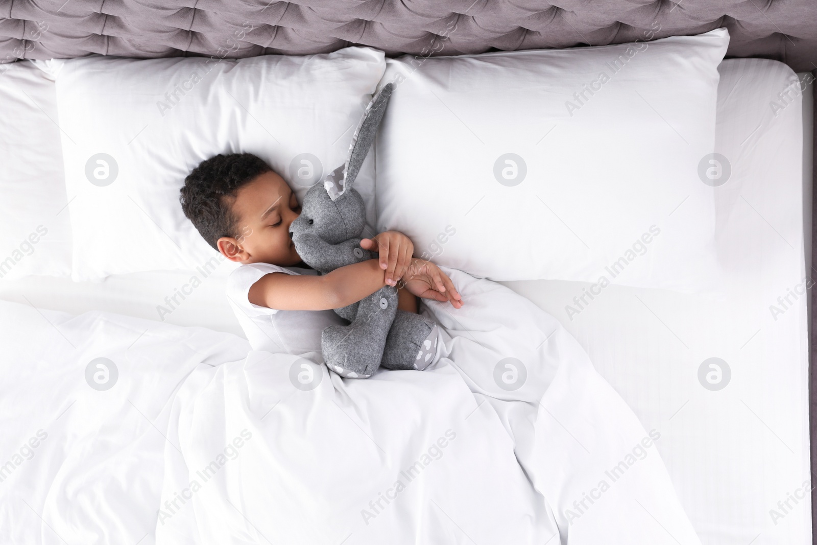 Photo of Cute little African-American boy with toy rabbit sleeping in bed, top view. Space for text
