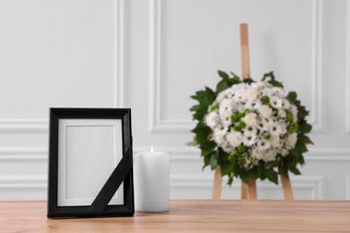 Photo frame with black ribbon, burning candle on table and wreath of flowers near white wall indoors, space for text. Funeral attributes