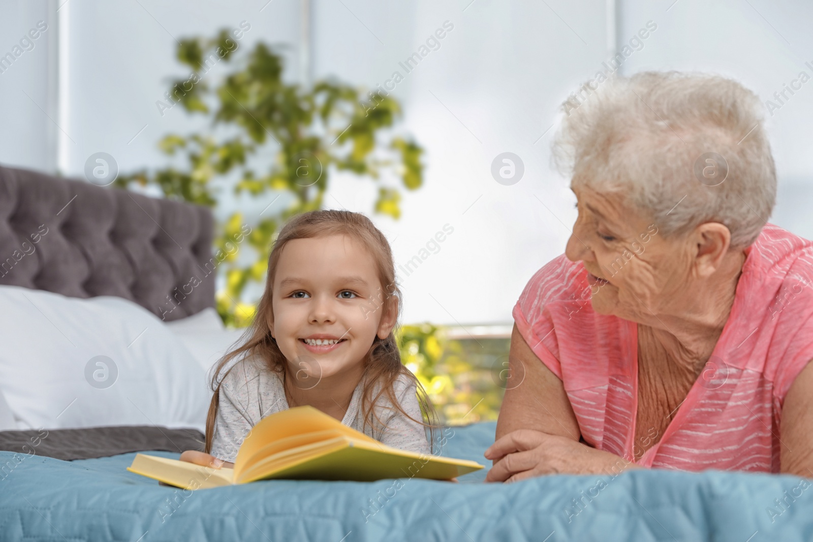 Photo of Cute girl and her grandmother reading book on bed at home