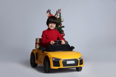 Photo of Cute little boy with Christmas tree and gift box driving children's electric toy car on white background