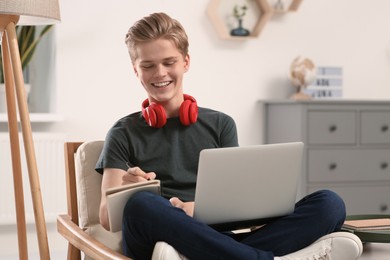 Photo of Online learning. Smiling teenage boy with laptop writing in notebook at home