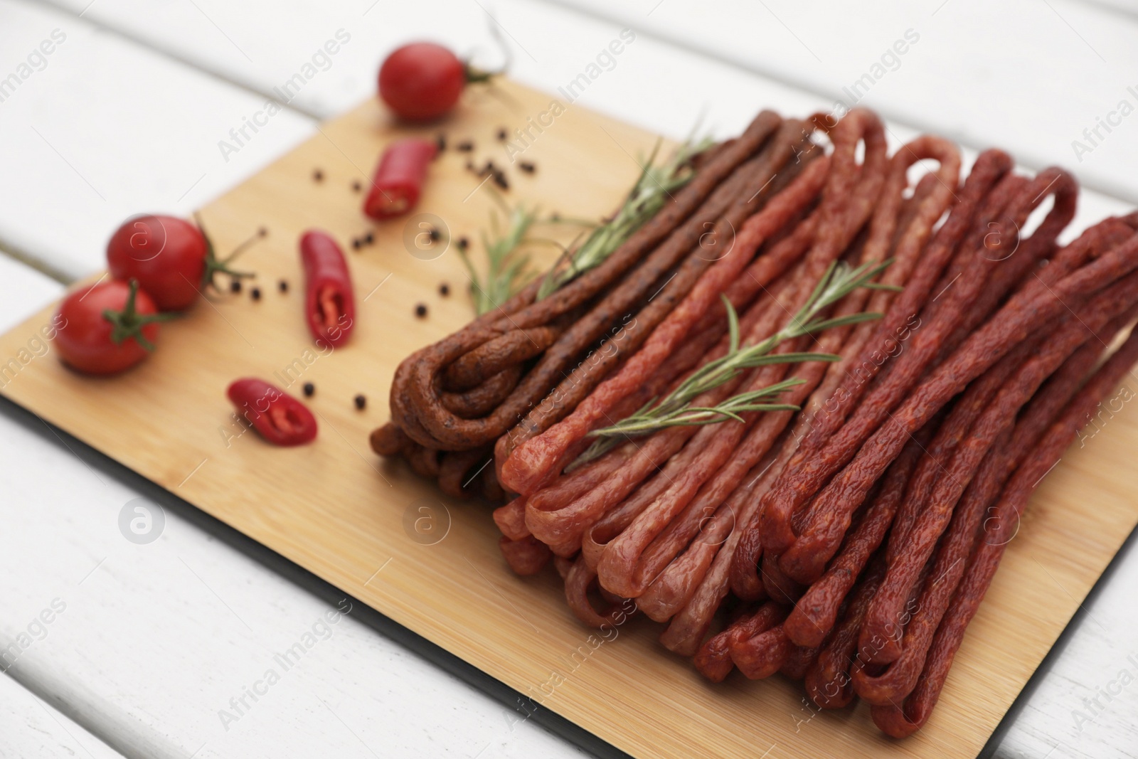 Photo of Delicious kabanosy with rosemary, peppercorn, chilli and tomatoes on white wooden table, closeup