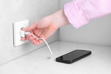 Photo of Woman plugging smartphone into power socket at white table indoors, closeup