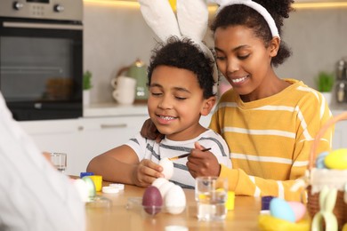 Photo of Happy African American mother and her cute son painting Easter eggs at table in kitchen