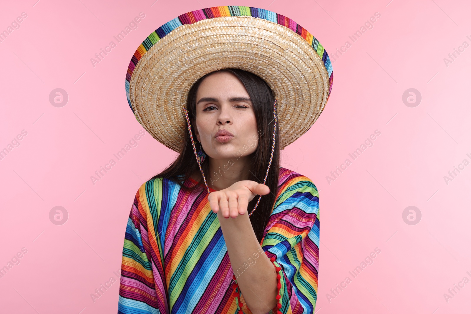 Photo of Young woman in Mexican sombrero hat and poncho blowing kiss on pink background