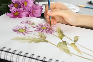 Photo of Woman drawing beautiful chrysanthemum flowers in sketchbook at table, closeup