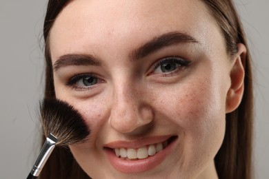 Smiling woman with freckles applying makeup with brush on grey background, closeup