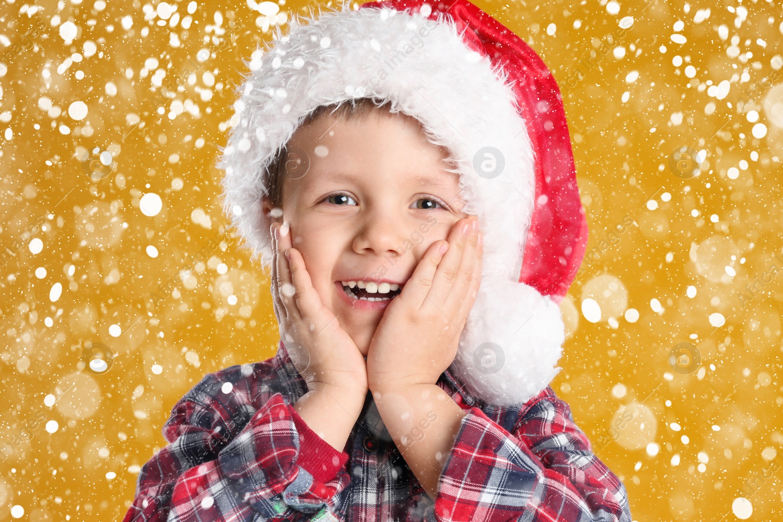 Image of Cute child in Santa hat under snowfall on yellow background. Christmas celebration
