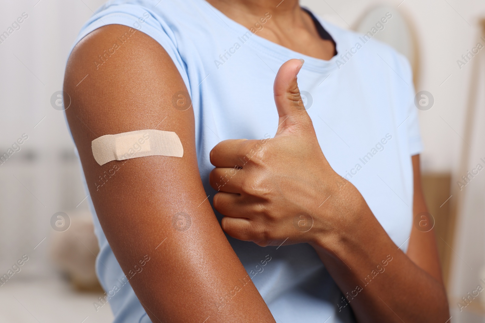 Photo of Young woman with adhesive bandage on her arm after vaccination showing thumb up indoors, closeup