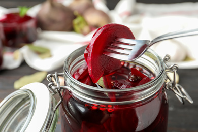 Photo of Fork with pickled beets over glass jar on wooden table, closeup