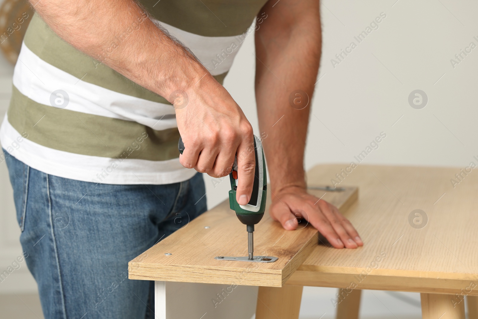 Photo of Man with electric screwdriver assembling furniture at table indoors, closeup