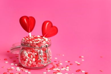 Sweet heart shaped lollipops and sprinkles in glass jar on pink background, space for text. Valentine's day celebration