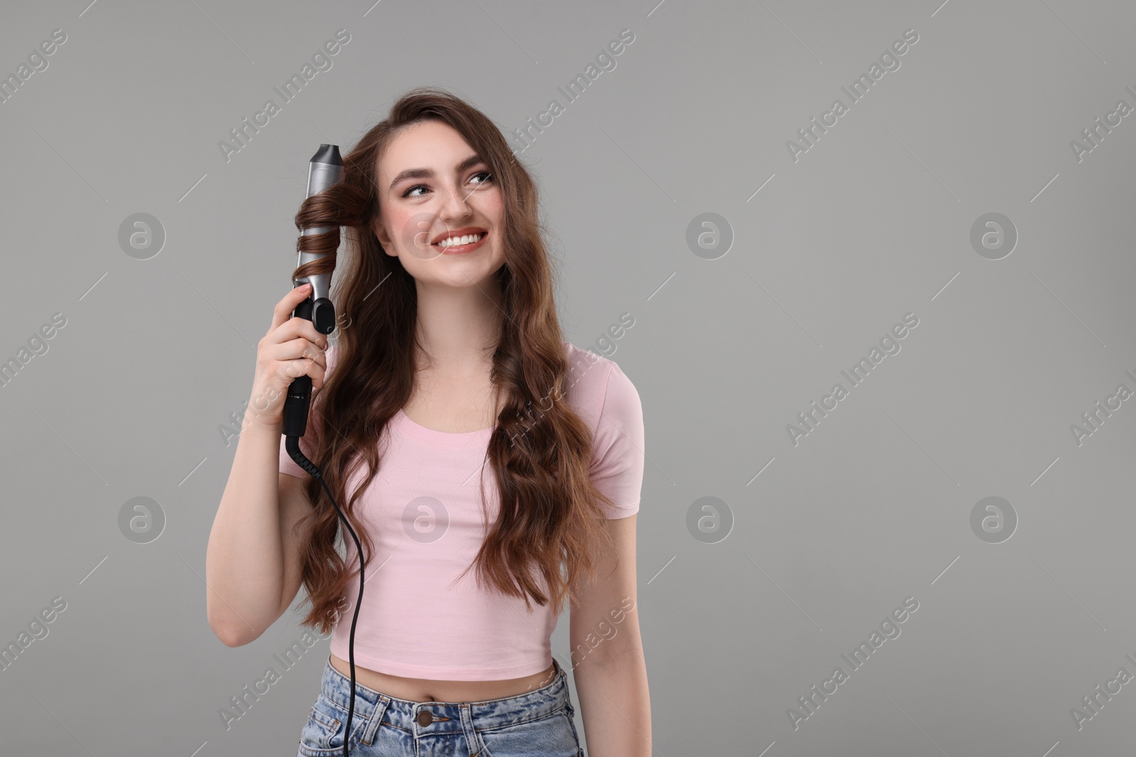 Photo of Beautiful young woman using curling hair iron on grey background, space for text