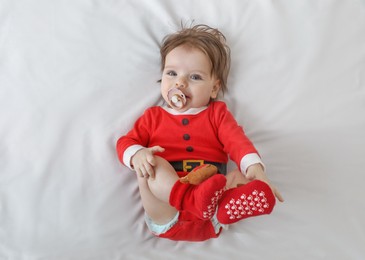 Cute baby wearing festive Christmas costume with pacifier on white bedsheet, top view