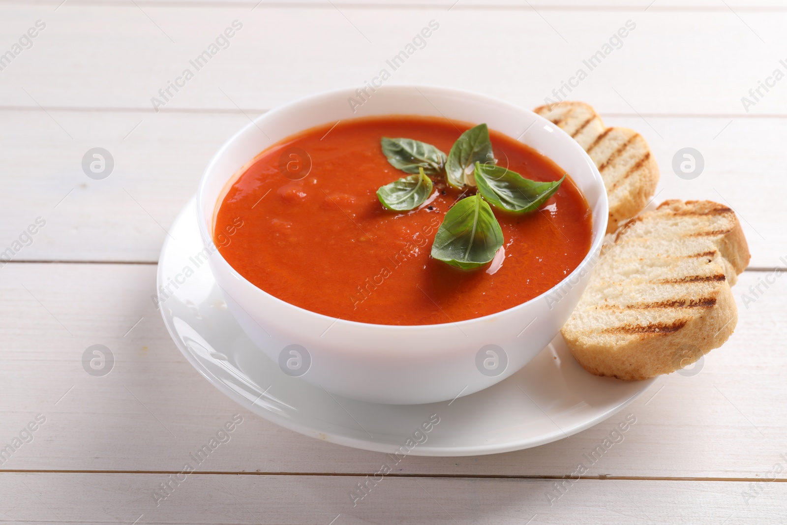 Photo of Delicious tomato cream soup in bowl with pieces of grilled bread on white wooden table