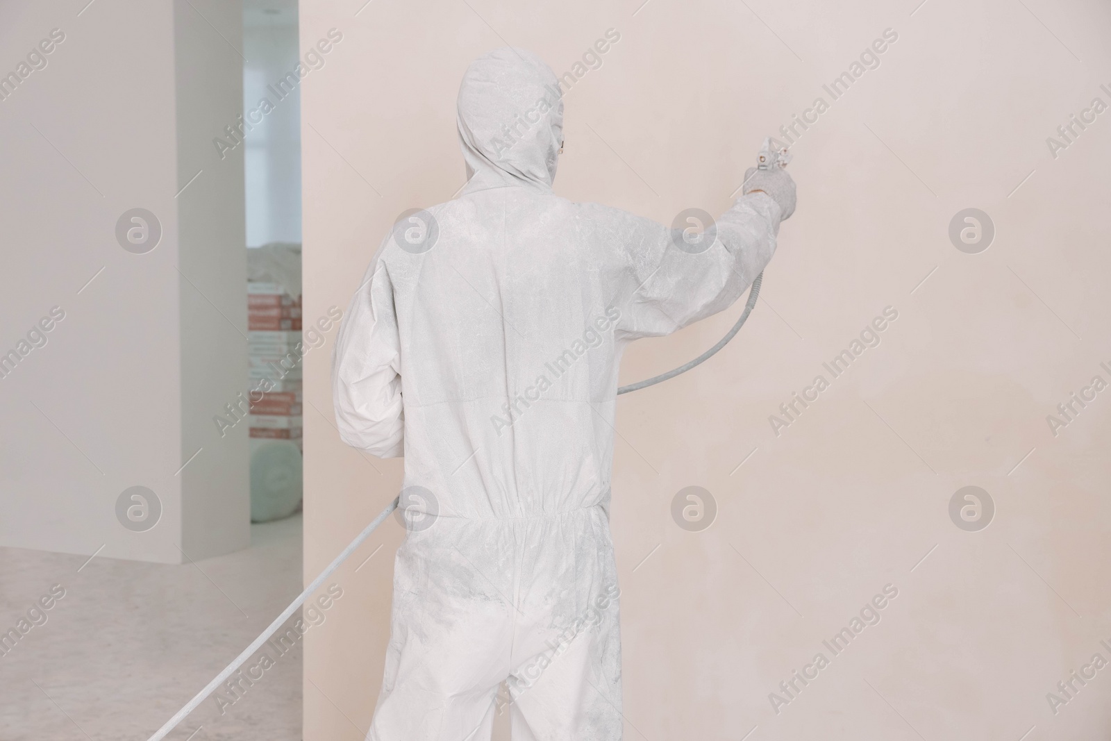 Photo of Decorator in uniform painting wall with sprayer indoors, back view