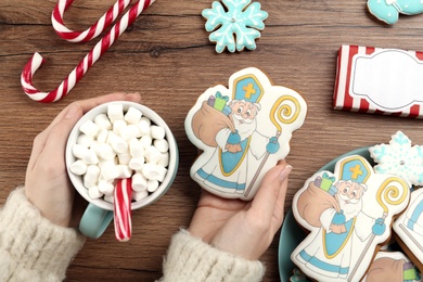 Photo of Woman with tasty gingerbread cookie and cup of hot drink at wooden table, top view. St. Nicholas Day celebration