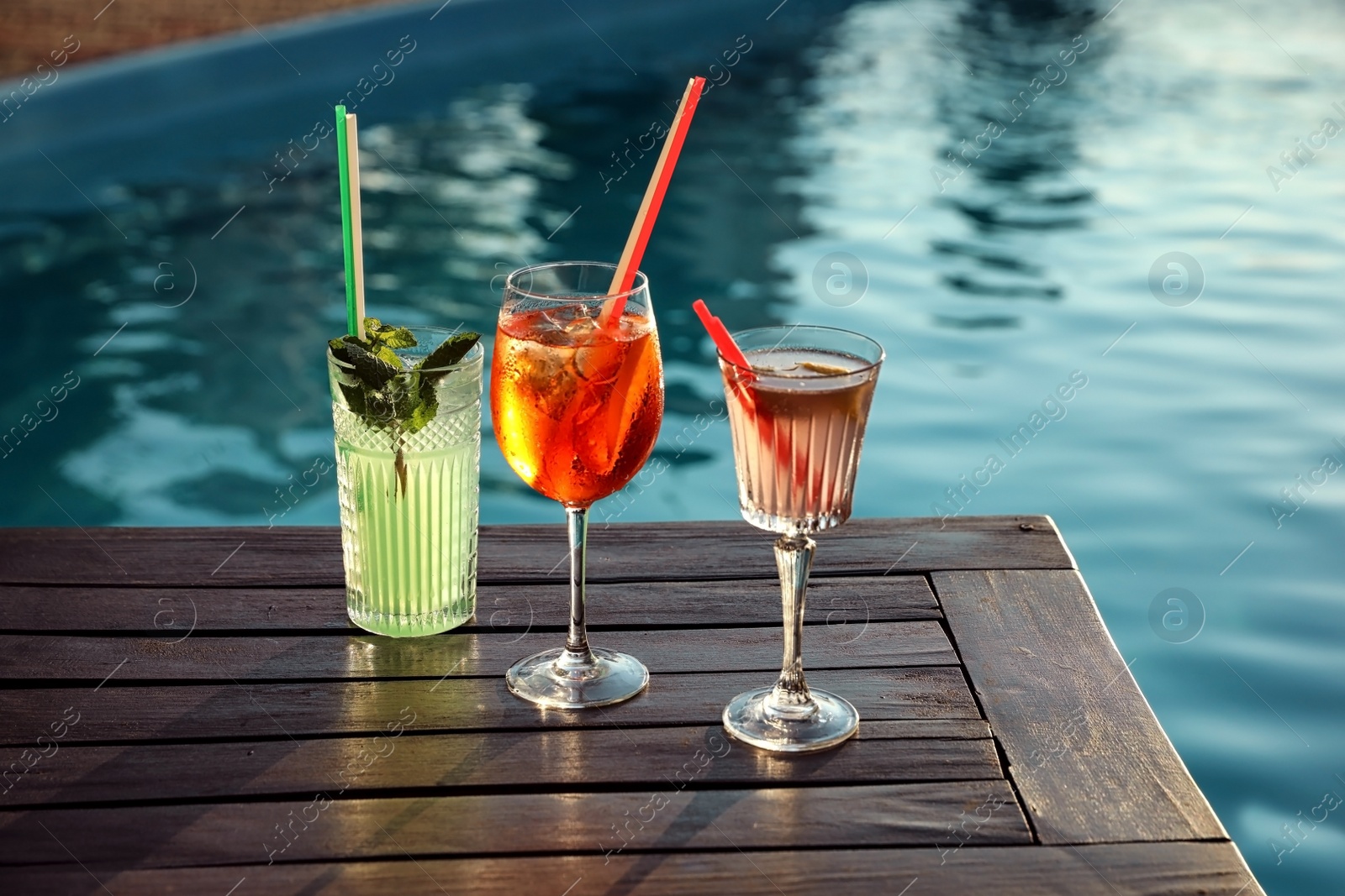 Photo of Glasses of fresh summer cocktails on wooden table near swimming pool outdoors