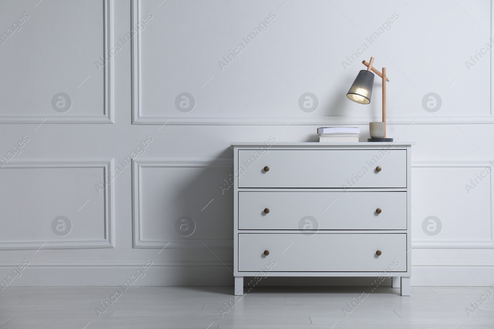 Photo of Lamp and books on chest of drawers near white wall indoors