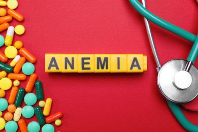 Photo of Word Anemia made with yellow cubes, pills and stethoscope on red background, flat lay